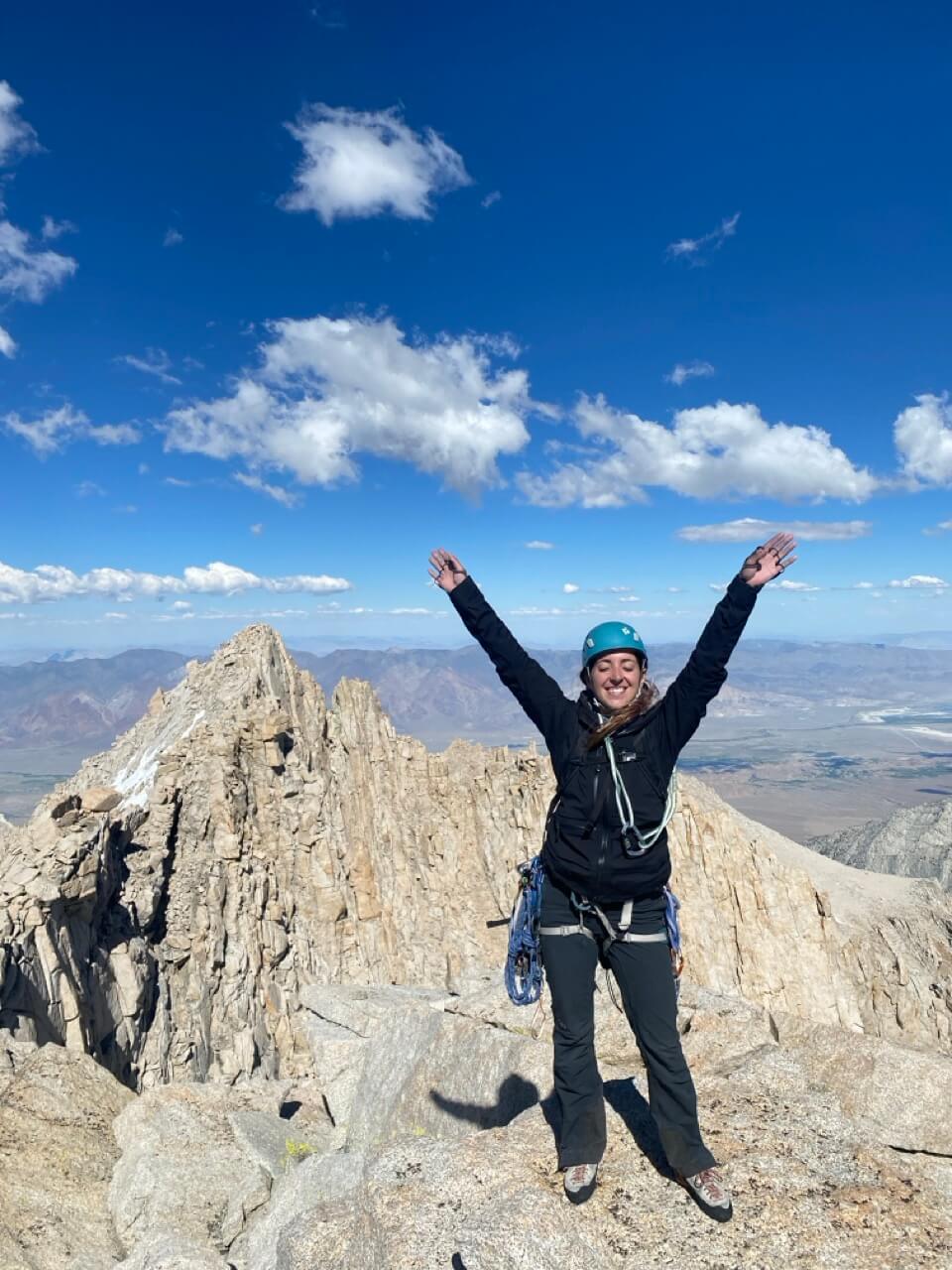 A woman standing a top a high peak. Her hand are in the air. 